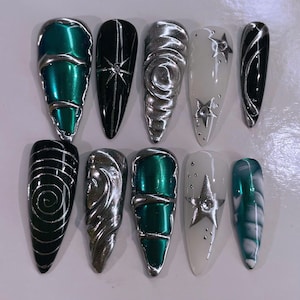 May include: Ten long, almond-shaped fake nails with different designs. The nails are painted in black, white, and green with silver accents. Some nails have a swirl design, others have stars, and some have a metallic finish.