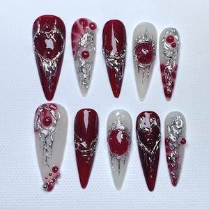 May include: A set of ten long, pointed artificial nails. The nails feature a deep red color with silver metallic accents. Some nails have a clear base with red and silver designs, while others are solid red.