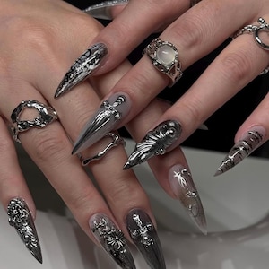 Puede incluir: Primer plano de uñas largas y puntiagudas, plateadas y grises, con varios diseños, incluyendo cruces, detalles florales y patrones abstractos. Varios anillos plateados con diseños únicos, como una piedra lunar y formas ornamentadas, también son visibles.