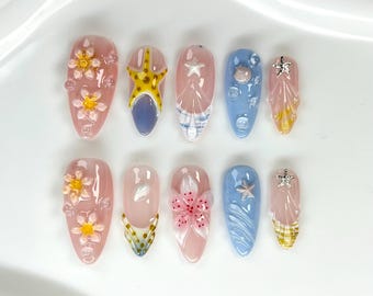 May include: A set of ten press-on nails featuring a variety of designs including pink bases with raised floral accents, blue bases with seashell and starfish charms, and clear tips with yellow and white striped patterns and silver starfish charms.