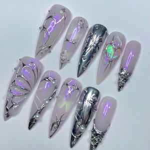 May include: A set of ten long, almond-shaped artificial nails. Each nail is painted a pale lavender color and adorned with silver metallic designs, including floral and abstract patterns, and small pearl accents. Some nails feature iridescent highlights.