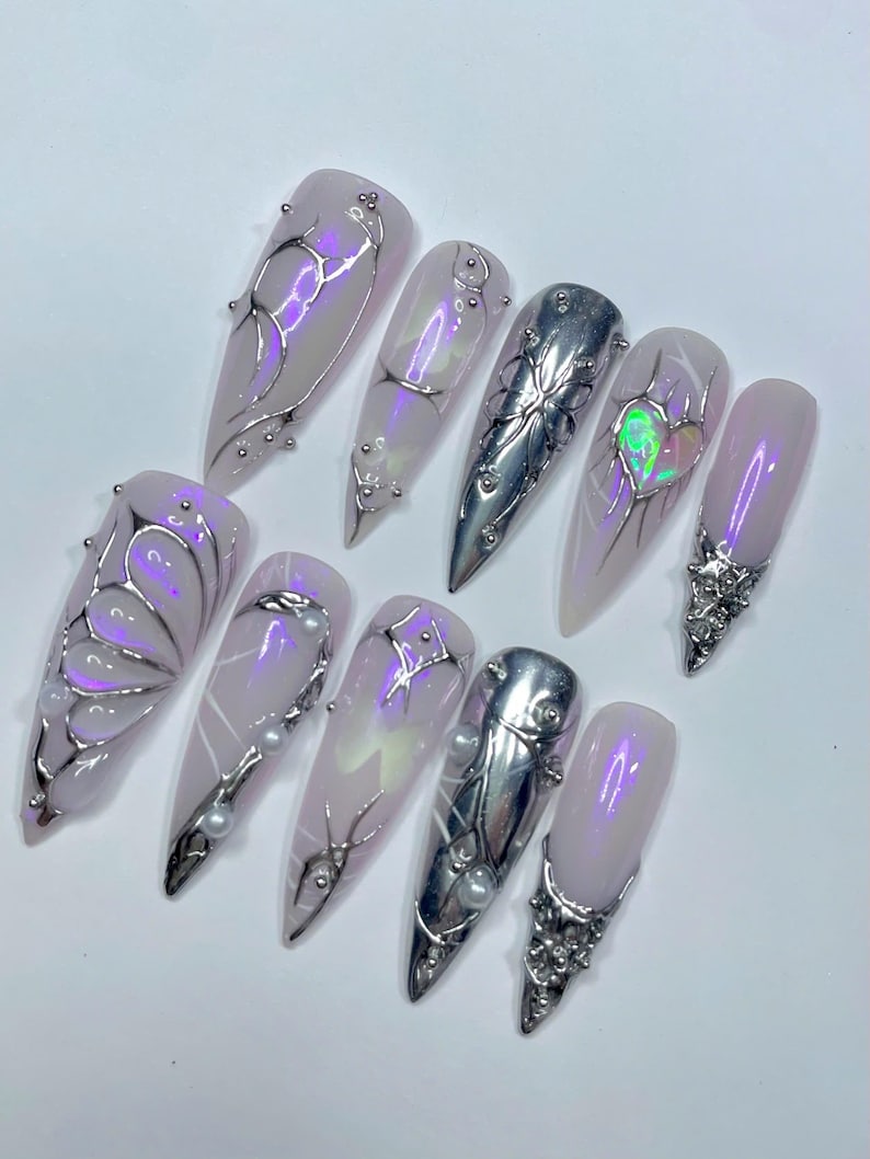 May include: A set of ten long, almond-shaped artificial nails. Each nail is painted a pale lavender color and adorned with silver metallic designs, including floral and abstract patterns, and small pearl accents. Some nails feature iridescent highlights.
