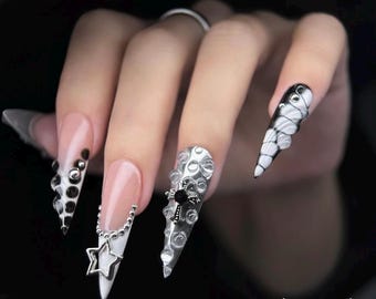 May include: A hand with long, pointed, artificial nails. The nails feature various designs, including black and silver embellishments, a star charm, and a black and white pattern. The nails are painted in a combination of white, pink, and silver.