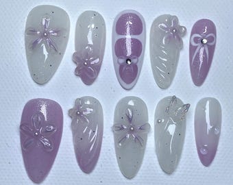 May include: Set of ten false nails with a light purple and white ombre design. Each nail features a different 3D flower design, including a bow, a swirl, and a flower with a pearl center.