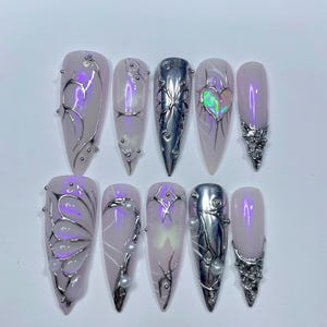 May include: A set of ten long, almond-shaped artificial nails. The nails are painted a pale pink with iridescent shimmer. Some nails feature silver metal designs, including butterfly and heart shapes, and small silver beads. One nail is solid silver.