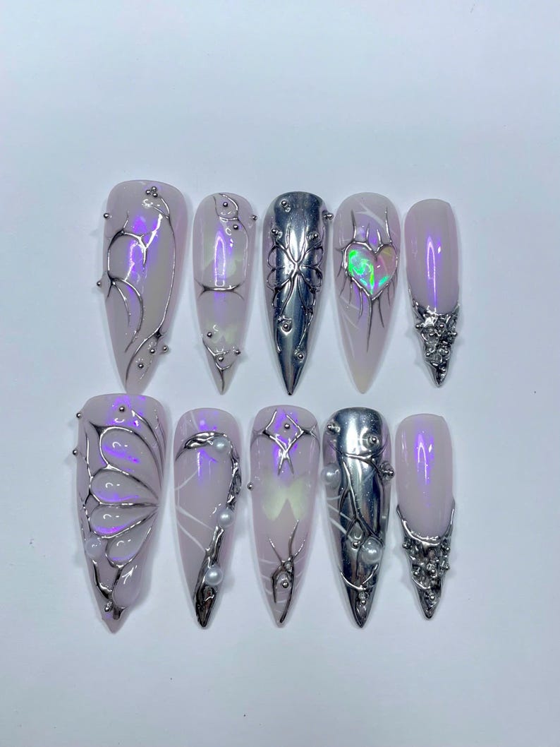 May include: A set of ten long, almond-shaped artificial nails. The nails are painted a pale pink with iridescent shimmer. Some nails feature silver metal designs, including butterfly and heart shapes, and small silver beads. One nail is solid silver.