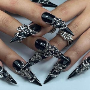K&ouml;nnte beinhalten: Schwarze, spitze Acryln&auml;gel mit silbernen Dekorringen. Die Ringe weisen verzierte Designs auf, darunter Kreuze, Perlen und florale Muster. Die N&auml;gel sind gl&auml;nzend und reichen &uuml;ber die Fingerspitzen hinaus.