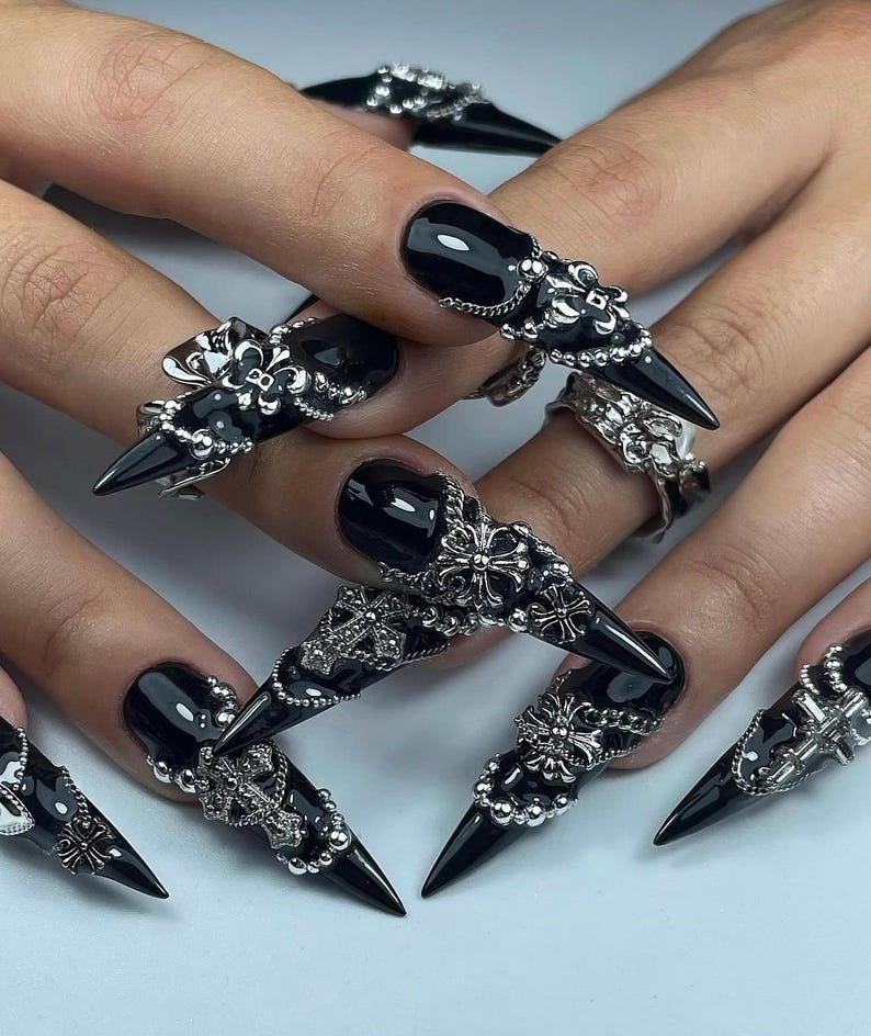 K&ouml;nnte beinhalten: Schwarze, spitze Acryln&auml;gel mit silbernen Dekorringen. Die Ringe weisen verzierte Designs auf, darunter Kreuze, Perlen und florale Muster. Die N&auml;gel sind gl&auml;nzend und reichen &uuml;ber die Fingerspitzen hinaus.