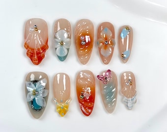 May include: A set of ten press-on nails featuring a nude base with orange and blue gradients. The designs include three-dimensional white and blue flower appliqués, a small pink butterfly charm, metallic gold and silver beads, and white pearls.