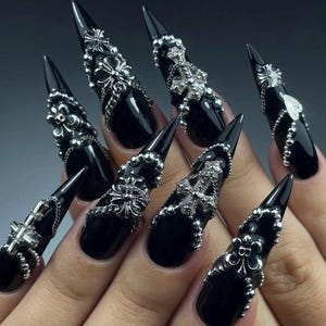 K&ouml;nnte beinhalten: Schwarze Stiletto-N&auml;gel mit silbernen Verzierungen. Jeder Nagel ist gl&auml;nzend schwarz lackiert und mit silbernen Verzierungen wie Kreuzen, floralen Designs und Perlen besetzt. Die N&auml;gel sind lang und spitz.