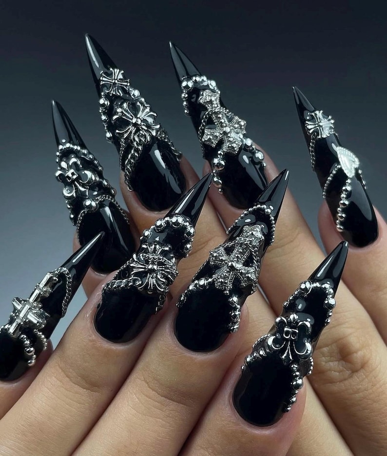K&ouml;nnte beinhalten: Schwarze Stiletto-N&auml;gel mit silbernen Verzierungen. Jeder Nagel ist gl&auml;nzend schwarz lackiert und mit silbernen Verzierungen wie Kreuzen, floralen Designs und Perlen besetzt. Die N&auml;gel sind lang und spitz.