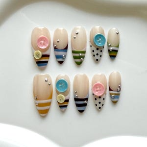 May include: A set of ten almond-shaped press-on nails featuring a nude base with various decorative elements. Designs include horizontal stripes in blue, yellow, brown, and green, black polka dots, small silver metallic studs, and pastel buttons.