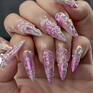 Op de afbeelding: Iriserende acrylnagels met een stiletto-vorm. De nagels hebben 3D bloemmotieven, parelaccenten en een wervelend, holografisch effect. Het kleurenpalet omvat tinten roze, paars en zilver, wat een glamoureuze uitstraling creëert.