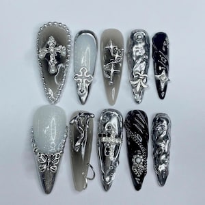 Puede incluir: Un conjunto de diez uñas artificiales largas en forma de almendra. Las uñas presentan una variedad de diseños, incluyendo cruces, estrellas y adornos metálicos abstractos. Los colores incluyen plata, negro y tonos de gris, con algunas uñas con acabado brillante.