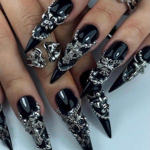 K&ouml;nnte beinhalten: Schwarze Stiletto-N&auml;gel mit aufwendiger silberner Nagelkunst und passenden Ringen. Die Nagelkunst zeigt Kreuze, Fleur-de-Lis und Perlenakzente. Die Ringe weisen &auml;hnliche Designs auf und schaffen eine zusammenh&auml;ngende gotische &Auml;sthetik.