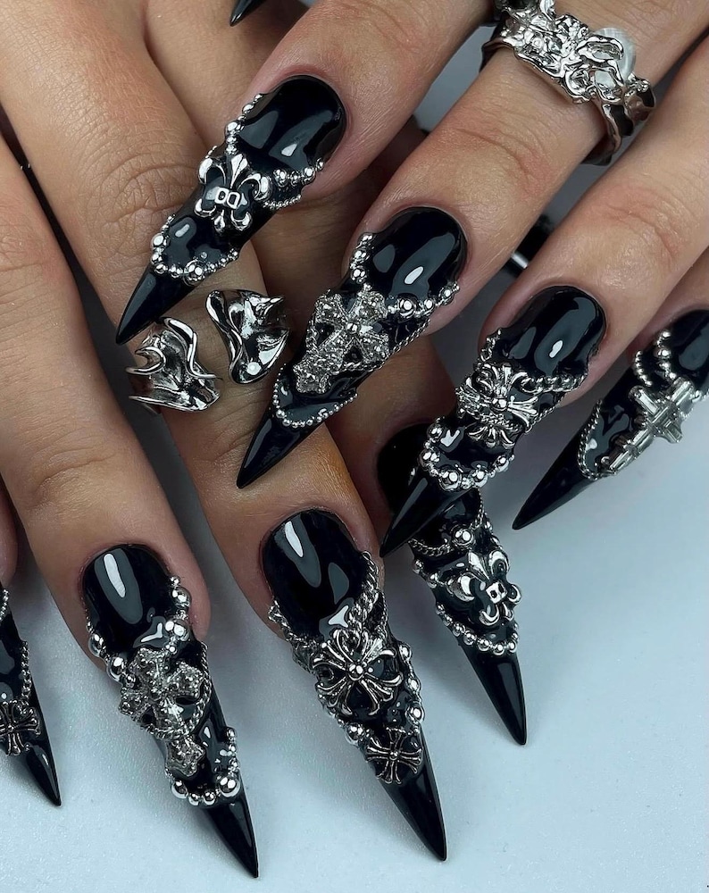 K&ouml;nnte beinhalten: Schwarze Stiletto-N&auml;gel mit aufwendiger silberner Nagelkunst und passenden Ringen. Die Nagelkunst zeigt Kreuze, Fleur-de-Lis und Perlenakzente. Die Ringe weisen &auml;hnliche Designs auf und schaffen eine zusammenh&auml;ngende gotische &Auml;sthetik.