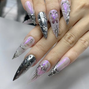 May include: A hand displaying long, pointed, artificial nails. The nails are painted with a pearlescent pink and adorned with silver metal designs, including floral and web-like patterns, and small pearl accents. Some nails feature a butterfly design.
