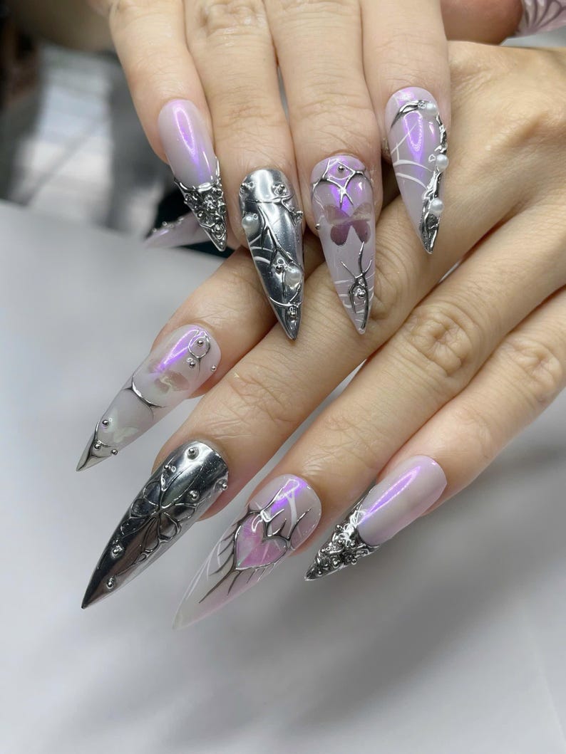 May include: A hand displaying long, pointed, artificial nails. The nails are painted with a pearlescent pink and adorned with silver metal designs, including floral and web-like patterns, and small pearl accents. Some nails feature a butterfly design.