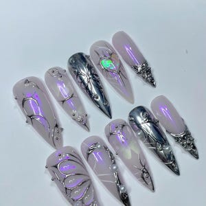 May include: A set of ten long, almond-shaped artificial nails. Each nail features a light pink base with silver and iridescent embellishments. Designs include metallic accents, pearls, and a heart-shaped iridescent gem.