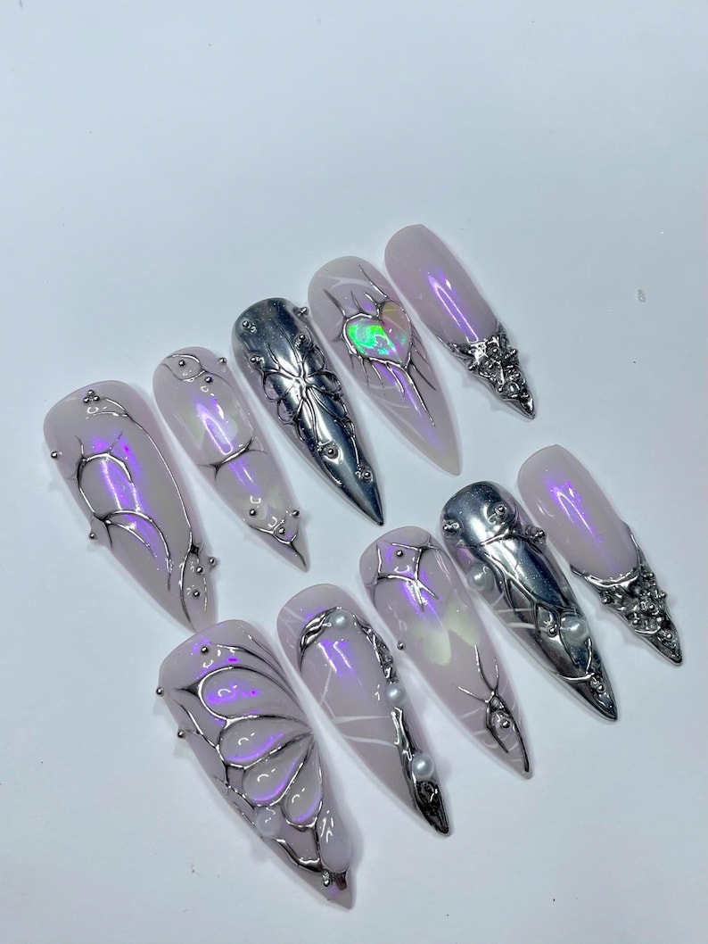 May include: A set of ten long, almond-shaped artificial nails. Each nail features a light pink base with silver and iridescent embellishments. Designs include metallic accents, pearls, and a heart-shaped iridescent gem.