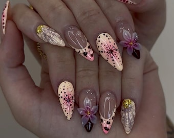 May include: A set of long, almond-shaped artificial nails. The nails feature a variety of designs, including pink and black polka dots, floral accents, and a French tip design. Some nails have 3D flower embellishments and gold accents.