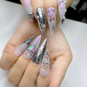 May include: A set of long, pointed fingernails with various designs. Some nails are silver-colored with pearl accents, while others feature a light purple hue with butterfly and abstract designs. The nails are adorned with metallic embellishments.