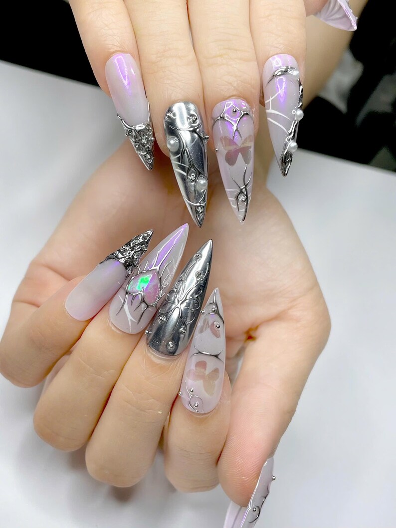 May include: A set of long, pointed fingernails with various designs. Some nails are silver-colored with pearl accents, while others feature a light purple hue with butterfly and abstract designs. The nails are adorned with metallic embellishments.