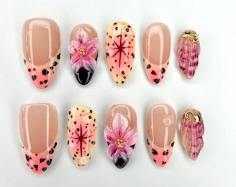 May include: A set of ten press-on nails featuring a nude base with various designs including pink floral accents, black polka dots, starburst patterns, and small gold star charms. The nails have a glossy finish and a mix of almond and oval shapes.