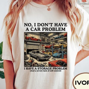 May include: An ivory short-sleeve t-shirt featuring a graphic print of a multi-level garage filled with various cars and a motorcycle. The text reads: NO, I DON'T HAVE A CAR PROBLEM I HAVE A STORAGE PROBLEM (And a severe lack of self-control).