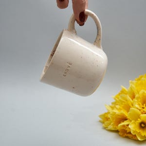 Puede incluir: Una taza de cerámica blanca con motas y la palabra "ERDVE" impresa en el lateral. La taza está siendo sostenida por una mano con esmalte de uñas rojo oscuro. La taza está enfocada, y el fondo es gris claro.