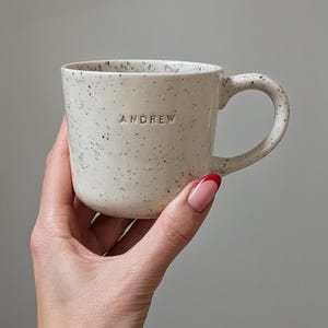 May include: A cream-coloured ceramic mug with a speckled finish and a rounded handle. The name ANDREW is embossed in capital letters on the front of the mug. The surface has a smooth texture with small dark flecks distributed across the body.