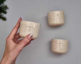 May include: Three small, speckled beige ceramic cups with a smooth finish. One cup is labeled MAMA with a small heart, one is labeled DADDY with a small heart, and the third features only a small heart. The cups have a cylindrical shape.
