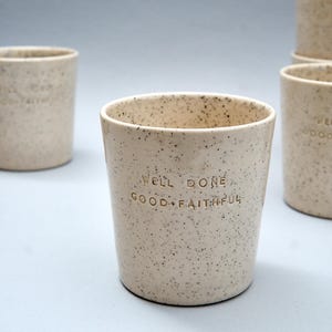 May include: A set of cream-colored ceramic cups with a speckled finish. Each cup features the embossed text WELL DONE GOOD + FAITHFUL on the front. The cups have a cylindrical shape with a smooth rim and a flat base, presented in a group.