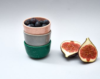 May include: Three speckled ceramic bowls stacked on top of each other. The top bowl is pink and contains blueberries. The bottom bowl is green. Two halves of a fig are on the white surface.