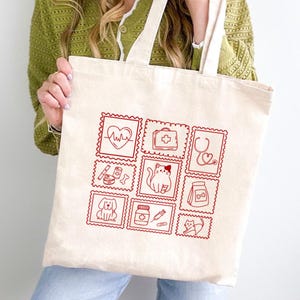 May include: A cream-coloured canvas tote bag featuring a grid of nine red line-art illustrations. The designs include a heart with an EKG line, a medical kit, a stethoscope, a dog, a cat, a medicine bottle, a syringe, and pet food supplies.