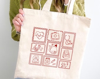 May include: A cream-colored canvas tote bag featuring a grid of nine red line-art illustrations. The designs include a heart with an EKG line, a medical kit, a stethoscope, a dog, a cat, a medicine bottle, a syringe, and pet food supplies.