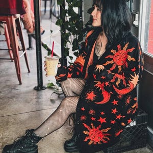 Könnte beinhalten: Eine schwarze Langarmstrickjacke mit einem leuchtend orangefarbenen und roten Himmelsmuster aus Sonnen, Monden und Sternen. Das Kleidungsstück wird über einem schwarzen Oberteil getragen, kombiniert mit einer schwarzen Netzstrumpfhose und Plateaustiefeln.