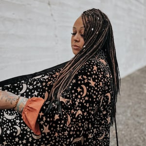 May include: A black velvet robe featuring a repeating pattern of gold crescent moons and stars. The garment has long sleeves with solid orange cuffs and a black trim along the front opening. The fabric has a soft texture and a celestial print.