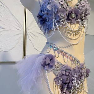 May include: A lavender two-piece set featuring a bra and matching bottoms adorned with fabric flowers, iridescent sequins, and cascading white pearl and silver rhinestone chains. The set includes sheer ruffled tulle accents on the side.