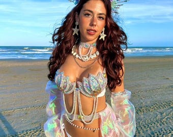 Puede incluir: Un conjunto de disfraz inspirado en sirenas que presenta un sujetador iridiscente en forma de concha y una braguita de bikini a juego adornada con hilos de perlas. El atuendo incluye mangas largas transparentes holográficas, una corona de conchas y accesorios para las piernas.