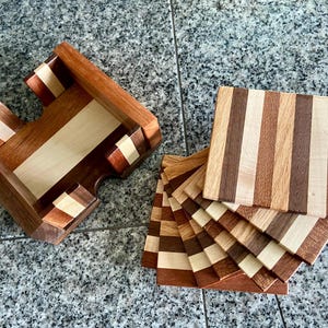 May include: A set of wooden coasters with a striped pattern in shades of brown, beige, and white. The coasters are stacked on top of each other and are displayed in a wooden holder.