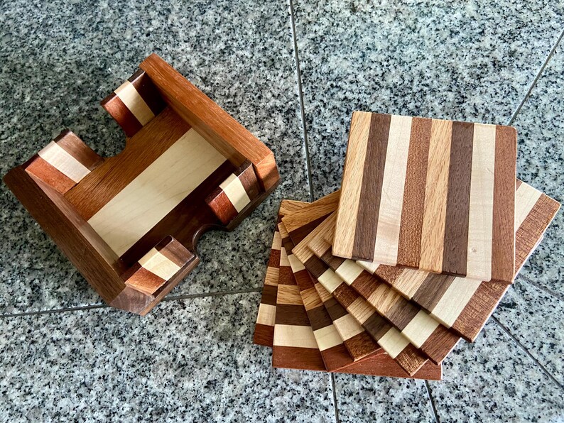 May include: A set of wooden coasters with a striped pattern in shades of brown, beige, and white. The coasters are stacked on top of each other and are displayed in a wooden holder.
