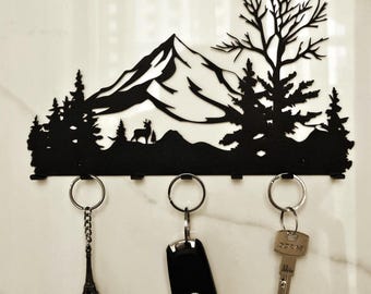 May include: Black metal key holder featuring a mountain and forest silhouette design. Includes three key rings with keys attached. The key holder has a decorative mountain and tree scene with a deer.