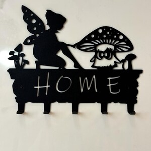 Può includere: Portachiavi in metallo nero con un design a forma di fata e fungo. Il portachiavi ha cinque ganci e la parola "HOME" è scritta sul davanti.