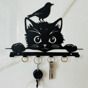 Puede incluir: Porta llaves de metal negro con forma de gato asomándose por un borde con un pájaro posado en su cabeza. El porta llaves tiene cinco ganchos para colgar llaves.