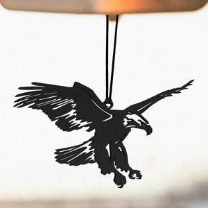 Può includere: Silhouette nera di un'aquila in volo, con le ali spiegate e gli artigli estesi. L'aquila è sospesa da un cordino nero, suggerendo che si tratti di un oggetto decorativo. Il design è dettagliato, catturando la forma potente dell'uccello.