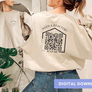 May include: Beige sweatshirt featuring a house graphic and the text "Need a Realtor? Scan Me" with a QR code. The front of the sweatshirt has a small house graphic and text. The words "Digital Download" are at the bottom.