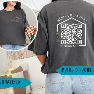 May include: Dark grey t-shirt with a white house logo and text on the front. The back of the shirt features a QR code within a house outline, with the text "Need a Realtor? Scan Me". The shirt is printed front and back.