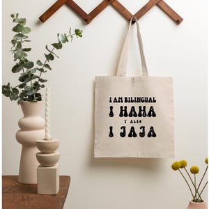 May include: A white canvas tote bag with black text that reads "I am bilingual I haha y also I jaja". The bag is hanging on a wooden wall rack.