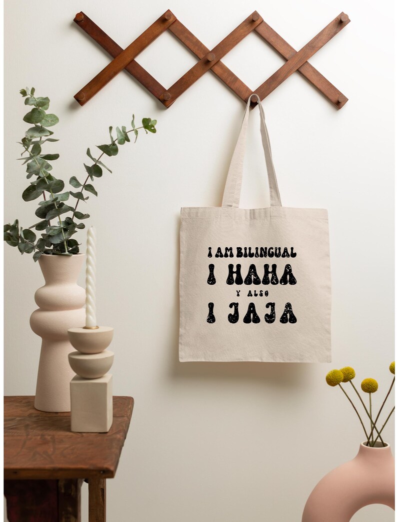 May include: A white canvas tote bag with black text that reads "I am bilingual I haha y also I jaja". The bag is hanging on a wooden wall rack.